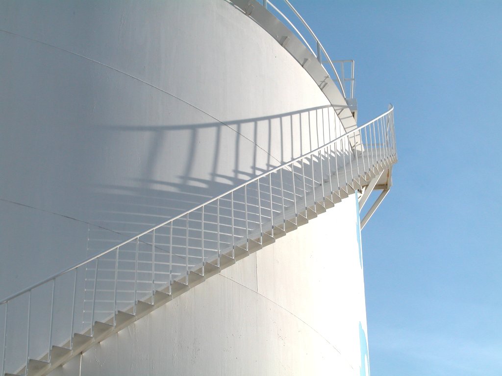Weiße Metalltreppe, die Schatten auf einen großen industriellen Speichertank unter klarem blauen Himmel wirft